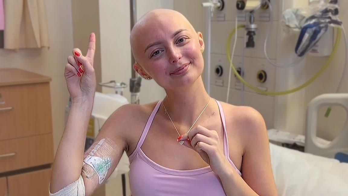 Person sitting in a hospital room wearing a pink top, with a bandaged arm and medical dressing visible. The person holds a necklace while raising one finger. Hospital equipment, tubing, and a bed are visible in the background.