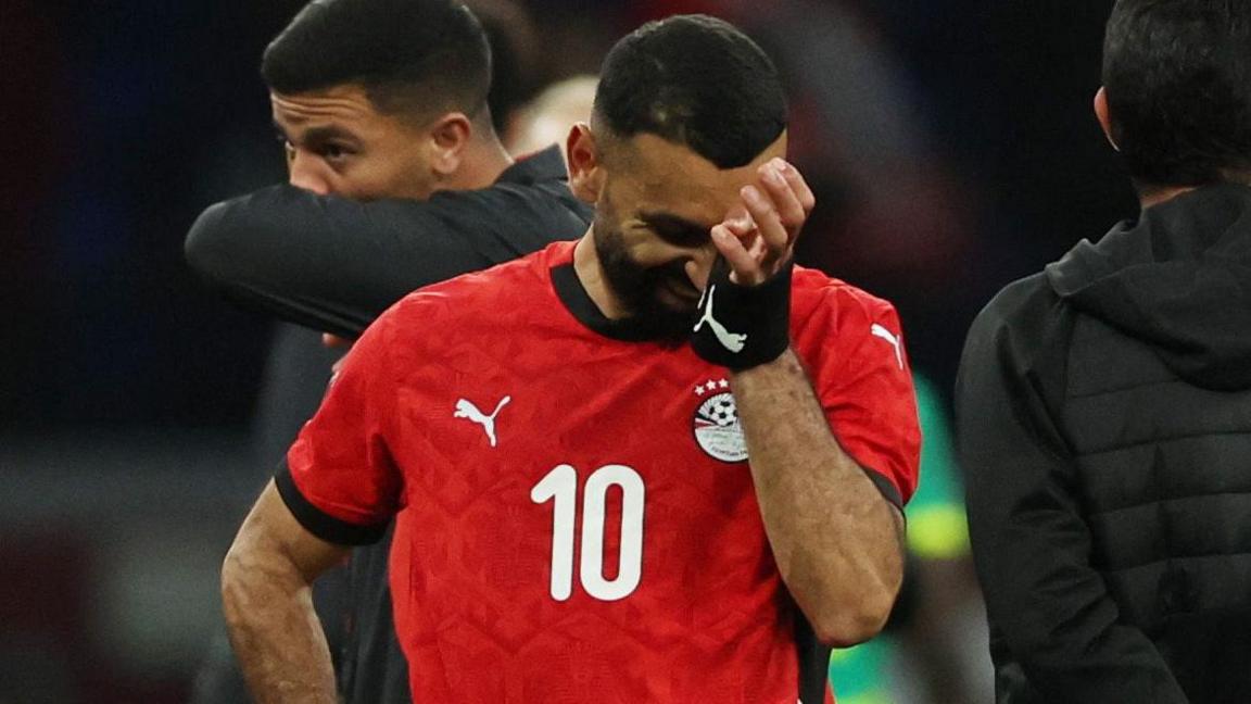 Mohamed Salah holds his left hand to his face as he looks downcast after Egypt's defeat by Senegal at the 2025 Africa Cup of Nations. He is wearing a red Egypt jersey with a white number 10 on the front and is seen from waist up. He is holding a black sweatband in the hand with which he is rubbing his face. Two other men dressed in black are seen behind Salah.