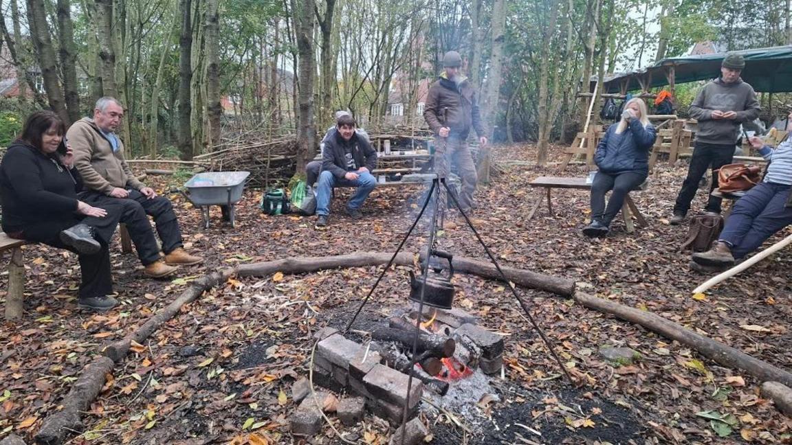 Two women and five men sat or stood in a circle in a wooded clearing, around a small wood fire with a metal frame hanging over it