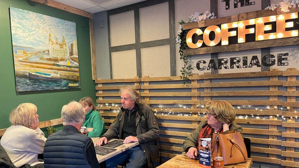 Customers in hte Coffee Carriage coffee shop. It has wooden palates on the wall - with COFFEE in lights above the tables. Some people have menus in front of them. A woman with short blonde hair sits next to a man with grey hair. A woman in a green fleece is opposite with a man in a wax jacket. On another table a woman with brown hair has a brown bag in front of her. she's wearing black rimmed glasses  and looking at the other table. 