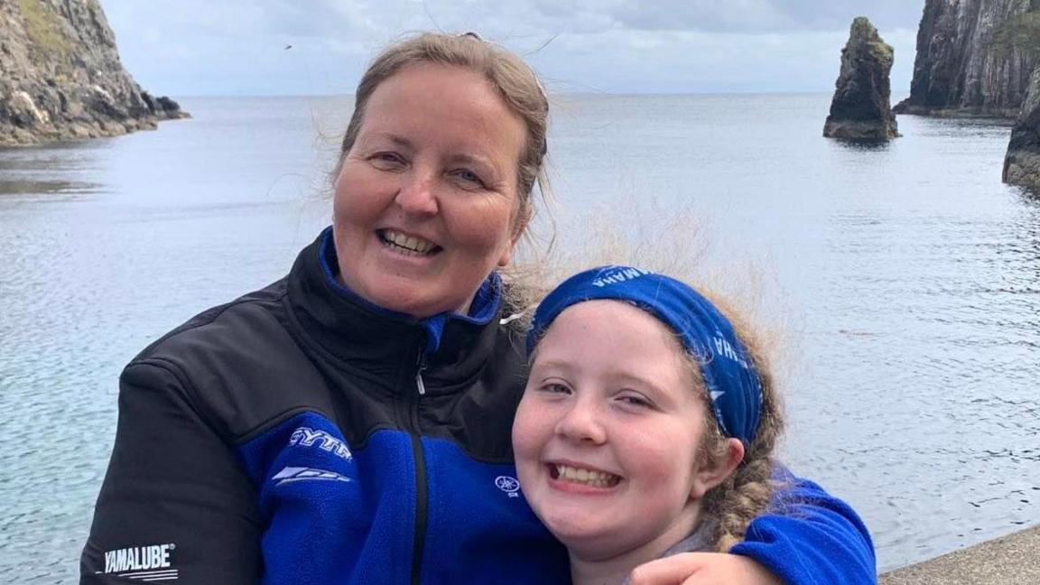 Heidi is hugging her daughter grace. She is wearing blue and black jacket, Grace is wearing a blue headband. The background is the sea with rocks coming out of it at both sides.