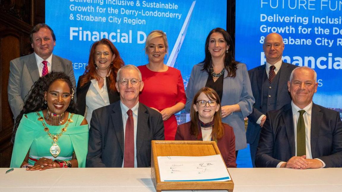 A delegation of ministers and public representatives, including Hilary Benn, Michelle O'Neill and Lilian Seenoi Barr, sit and stand behind a signed city deal 