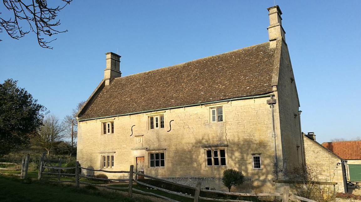 A two floor old house with yellow stone and a brown roof. A fence is located in front of it and greenery and trees can be seen to the left. There are three windows at the top and three at the bottom.