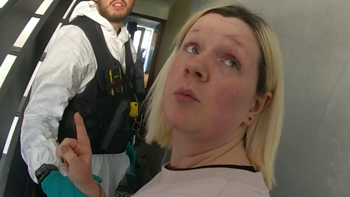 A picture from a body-worn video captured by a police officer. Virginia McCullough is looking over her left shoulder at the officer with a camera. She is wearing a faded pink sweater and being held by a second police officer wearing green gloves, a white forensic suit and black body armour.