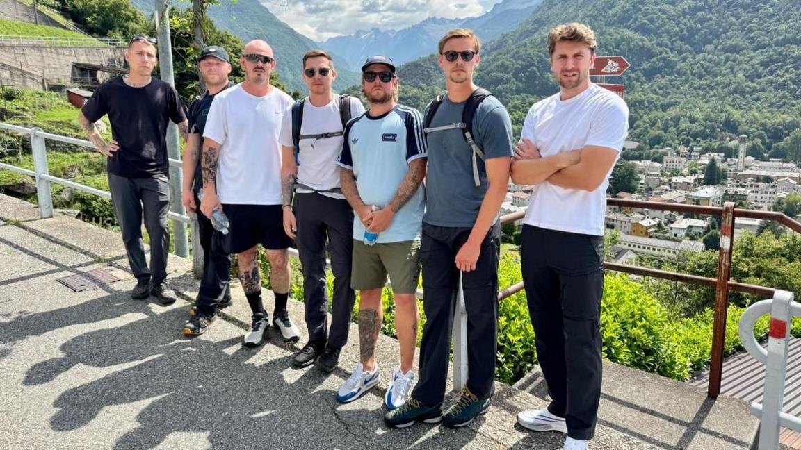 A group of seven men are standing in front of a metal fence which looks out on lots of buildings, trees and mountains below. Some are wearing backbacks and holding water bottles with serious expressions on their faces. The sun is out and it looks like a hot day.
