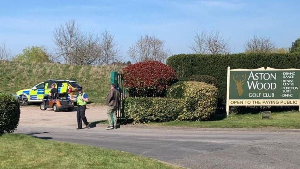 Green hedges are either side of a road and near to a police officer and a man with his back to the camera wearing a brown jacket and green trousers. A police car is parked on the left of the photo.