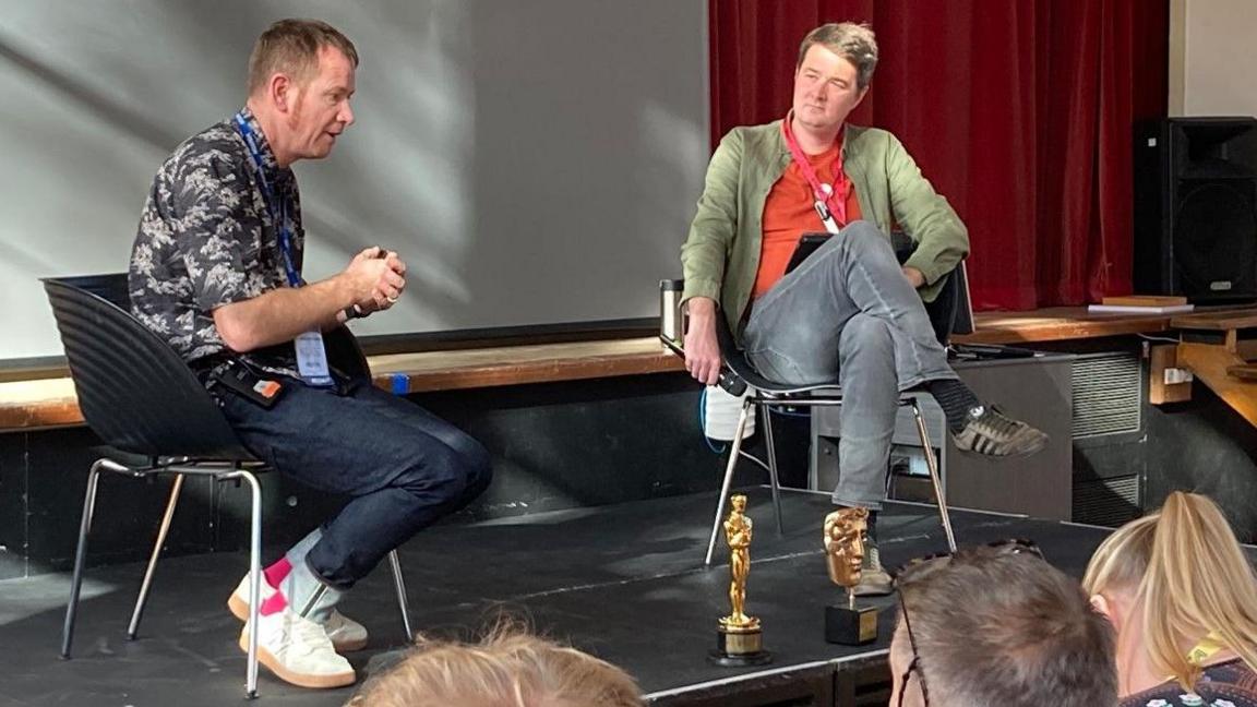 Two men sitting on chairs on a stage. The man on the left is wearing a dark patterned shirt with the sleeves rolled up, blue jeans, and white shoes. He has his hands clasped together and appears to be mid-speech. A man on the right holding a microphone looks towards the other man. On the front of the stage is an Oscar trophy and a Bafta trophy