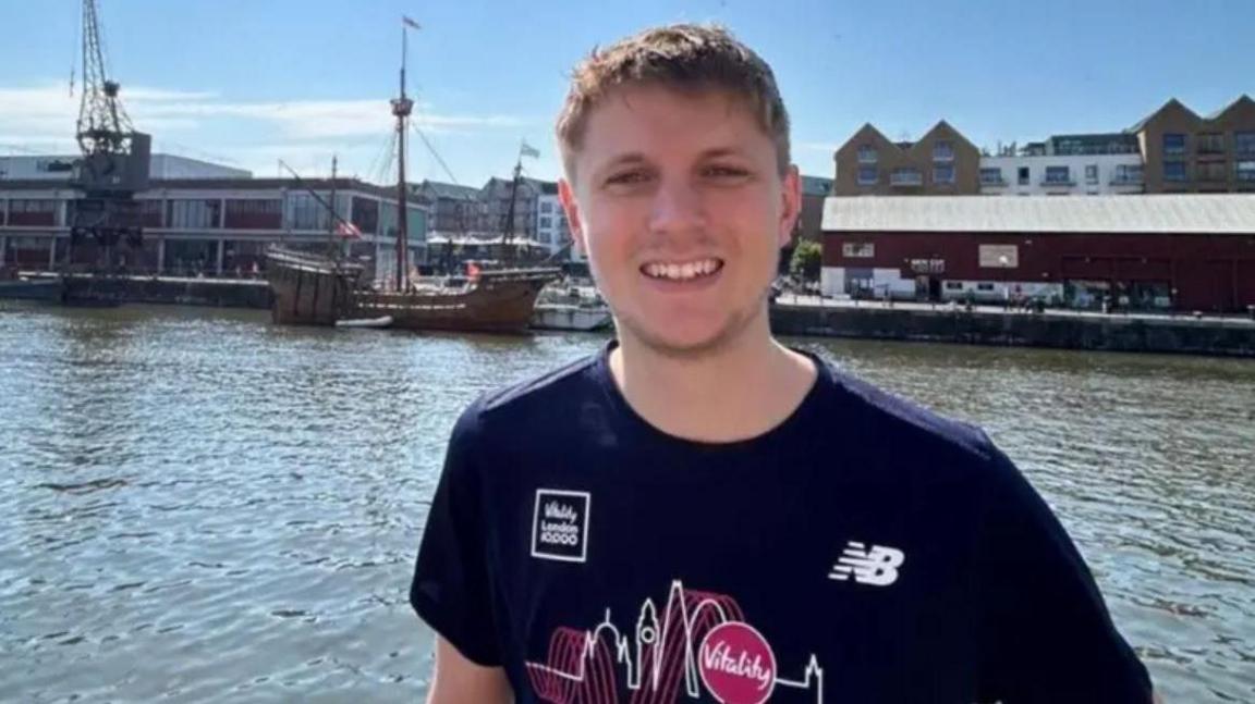 A head and shoulders shot of a man smiling at the camera wearing a navy T-shirt with a design on. He has short brown hair. Behind him is a river, boats and buildings in the distance. The sky is blue.