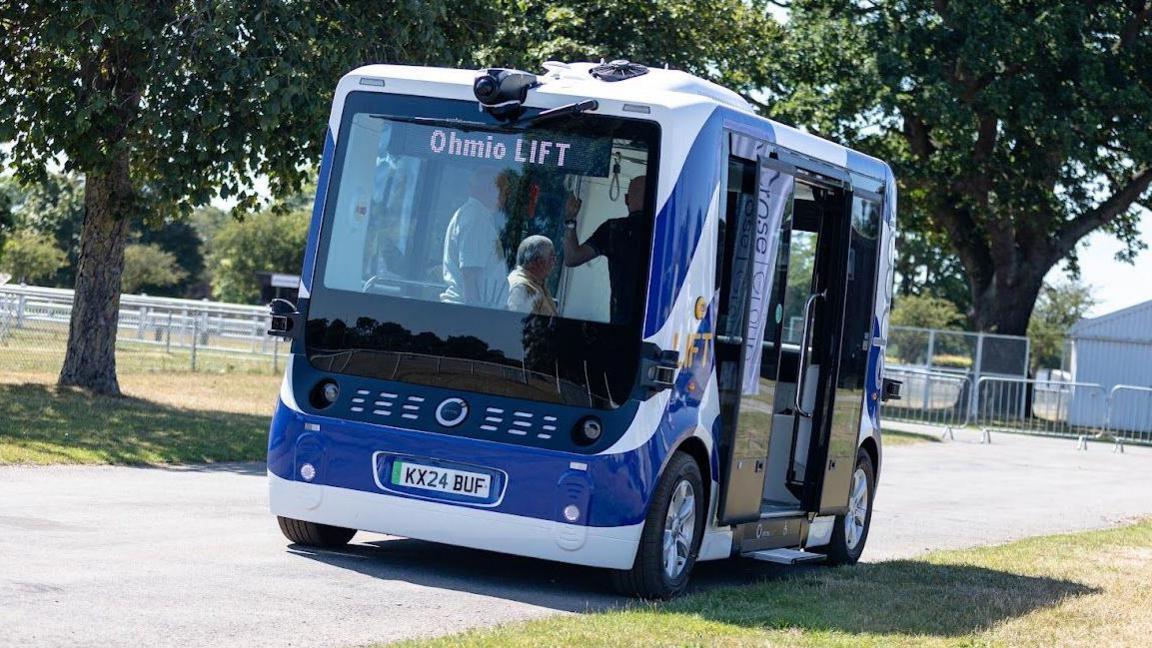 A view of the front of the prototype self-driving shuttle vehicle as it moves along a road. It is blue and white, has four wheels and large passenger doors. Passengers can be seen inside the vehicle.