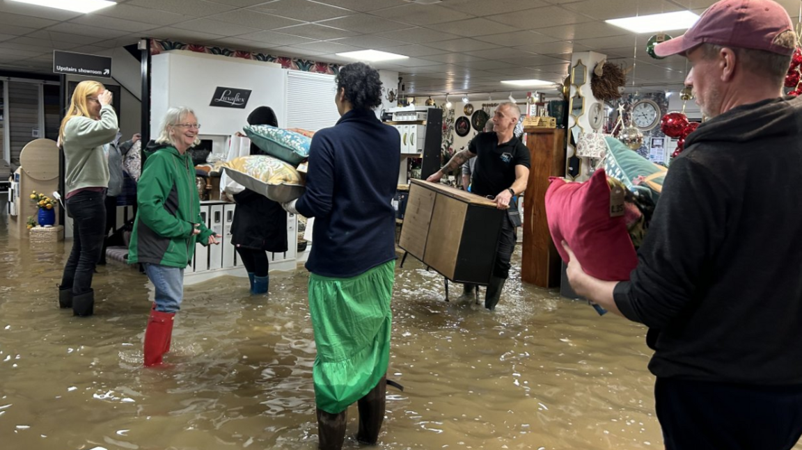 Staff inside a flooded carpet store, wearing wellies and carrying furniture 