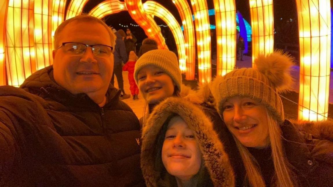 A family of four, a mother, father and two teenage girls, pose for a photo inside a light tunnel. It is winter and they are wearing bobble hats and fur-lined hoods. 