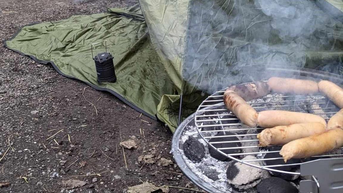 Sausages are cooking on a small barbecue with smoke rising off it. There is a green camping tent in the background.