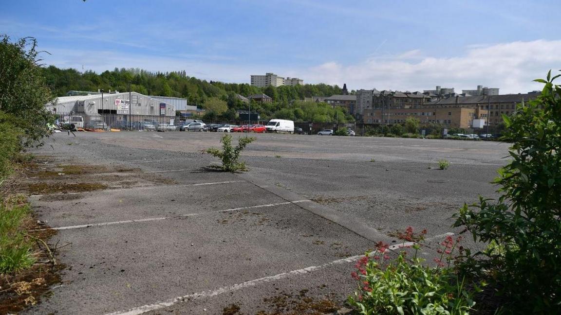 A derelict car park with weeds growing up through the tarmac. In the background is an industrial unit and blocks of flats.