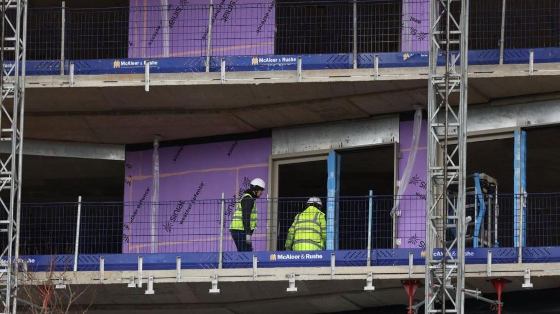 Two construction workers in hi-vis and hard hats on a balcony at a building site for residential properties in London. They are surrounded by metal scaffolding and purple insulation