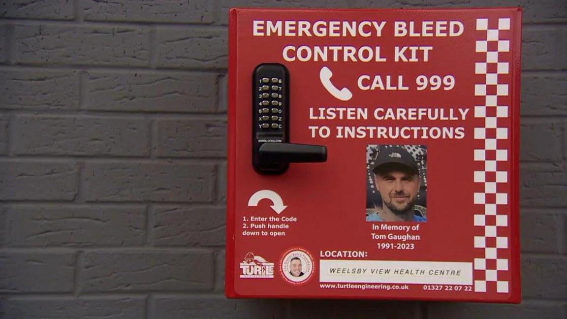 A red bleed kit box. It includes a black and silver key code, a photo of Tom Gaughan and white writing that reads: "Emergency bleed control kit. Call 999. Listen carefully to instructions. In memory of Ton Gaughan, 1991-2023."