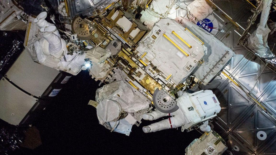 Butch and Suni float amidst highly technical equipment outside of the ISS, wearing white space suits. You can't tell who is who as one has their back to the camera, the other has their feet in the air. 