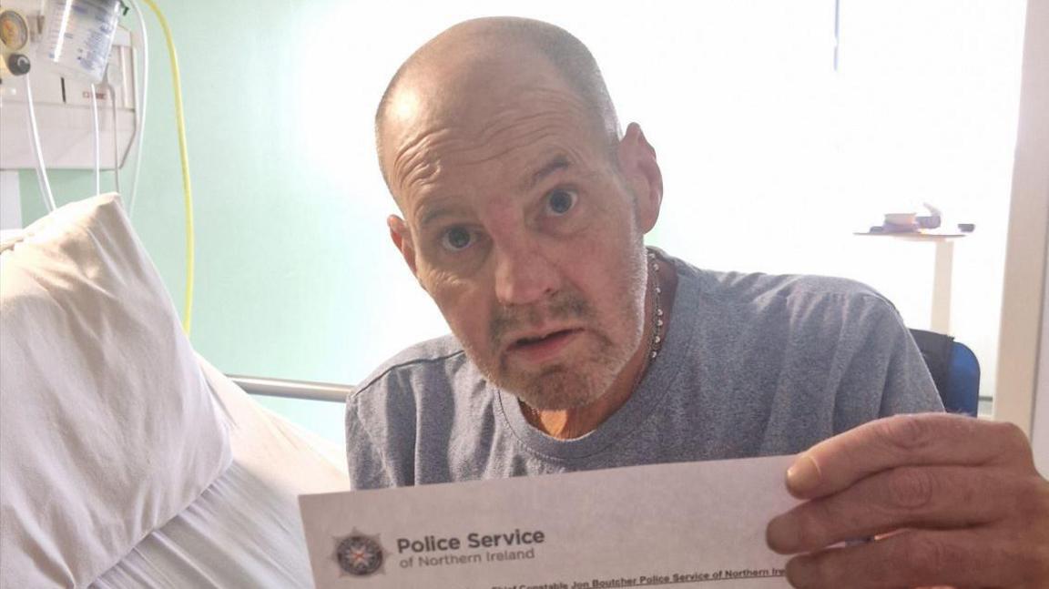 Eugene Thompson, with grey shirt on hospital bed, holding up a letter with a Police Service of Northern Ireland (PSNI) crest at the top left.