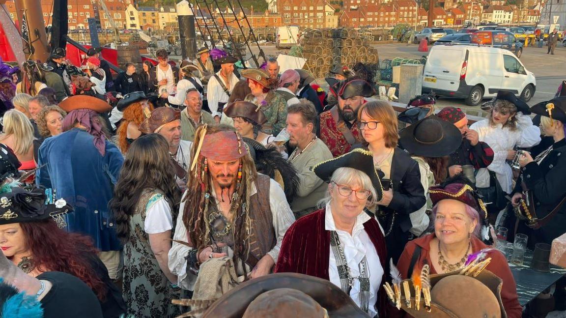 A group of people dressed like pirates at a show presented by the Auckland Shanty Singers. The singers are mingling with the crowd.