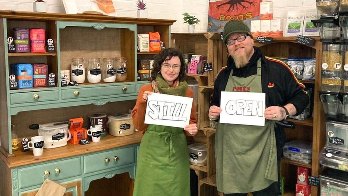 Two staff members at Roots refill standing in the shop holding signs that say "Still" and "open"