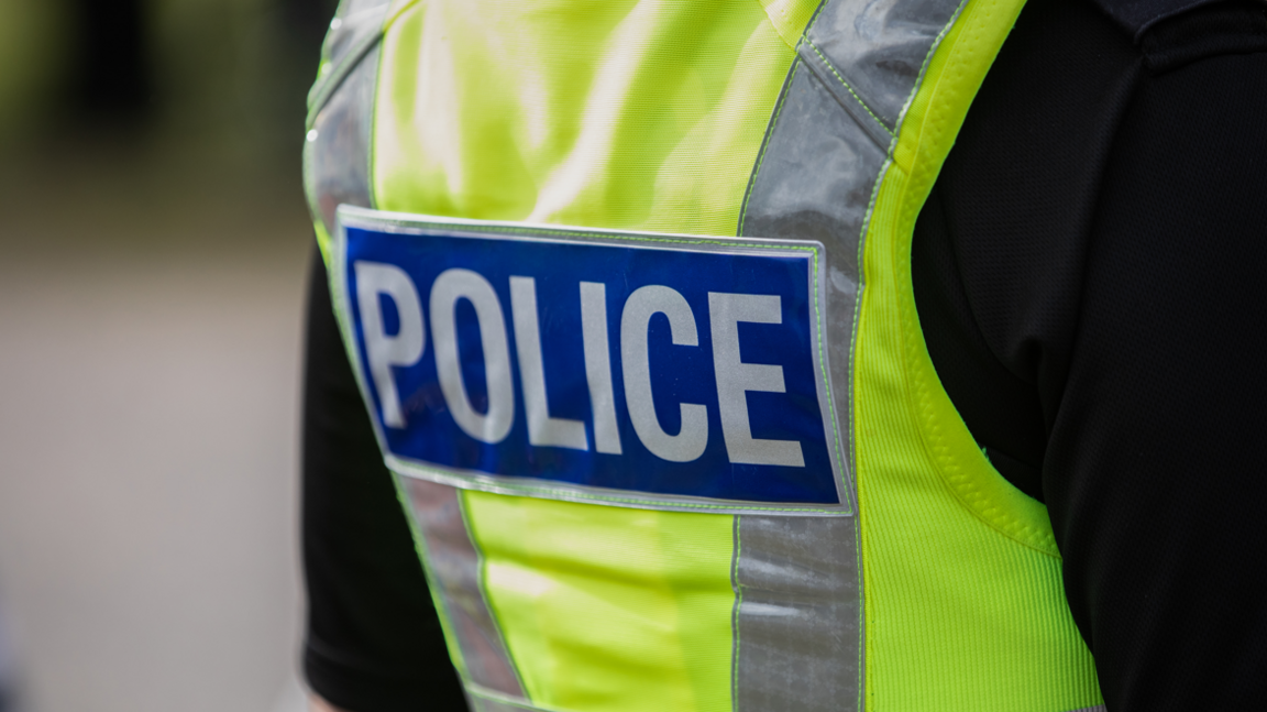 A close-up of the back of a police officer, who is wearing a uniform: a green hi-vis vest with police on the back in a blue rectangular square