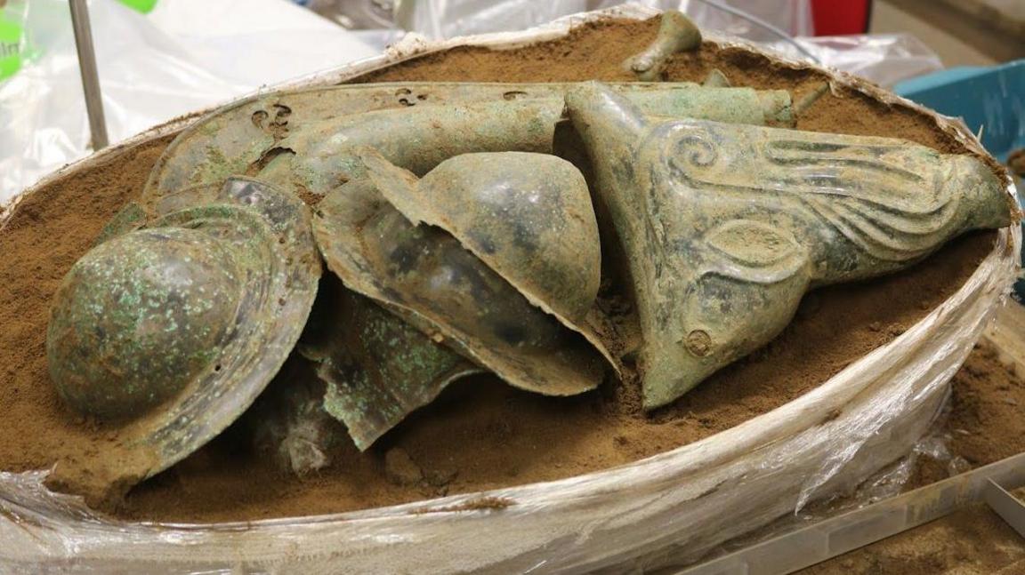 Hoard of Iron Age finds appearing from a block of sandy soil in a lab. Only the top layer can be seen, showing three shield bosses in the middle and on the right a boar's head, 