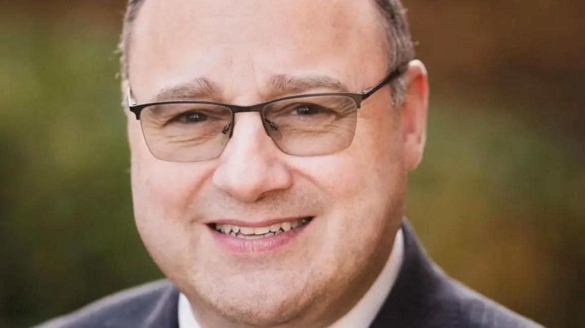 Close-up of Peter Reid, who has short brown hair and is wearing glasses and a grey suit jacket.