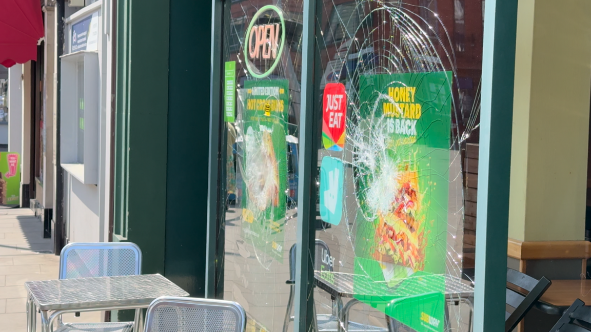 Another shop front window that has been smashed. Tables and chairs have been placed outside the business.