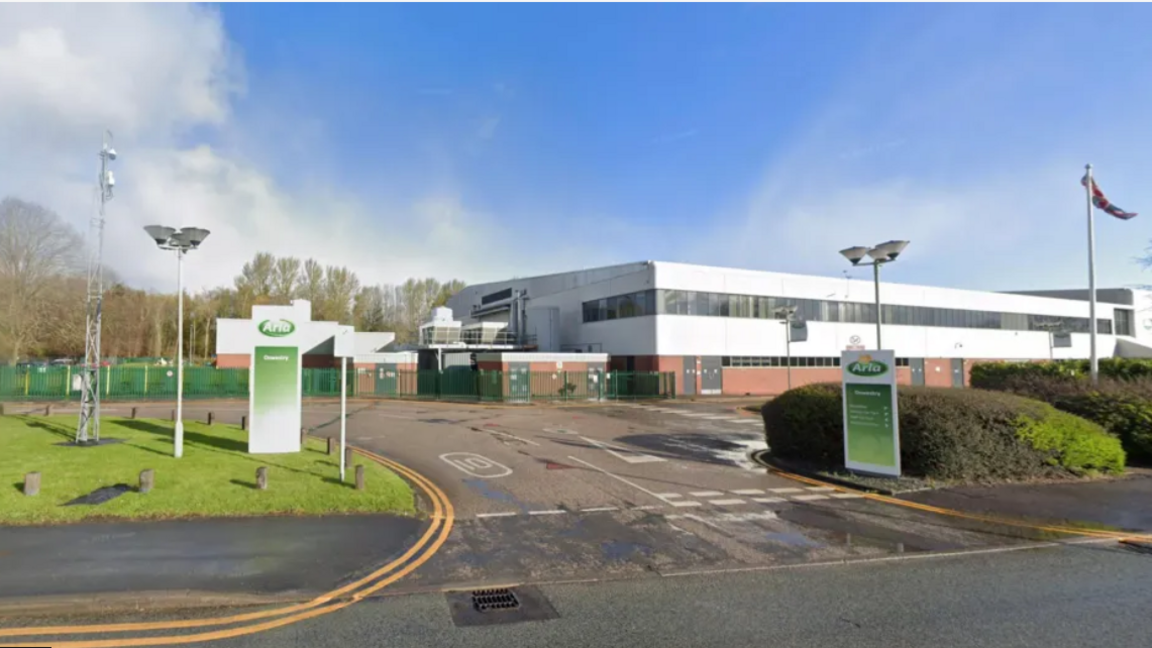 Arla Foods packaging plant showing an entrance off a road to a white and brick building behind signs saying "Arla"