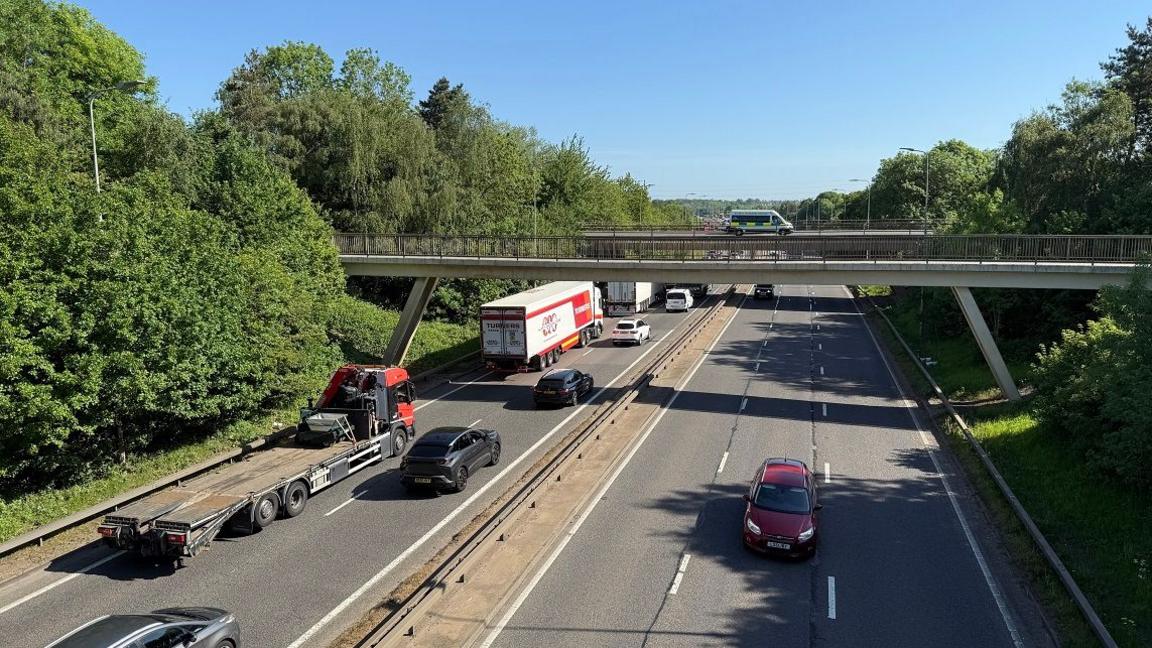 Dual carriageway showing traffic including lorries, vans and cars heading up from the bottom left of the photo. The vehicles are going under two motorway bridges. A police van is parked on the most distant bridge. A red van is visible heading in the opposite direction. There are trees on either side of the road.