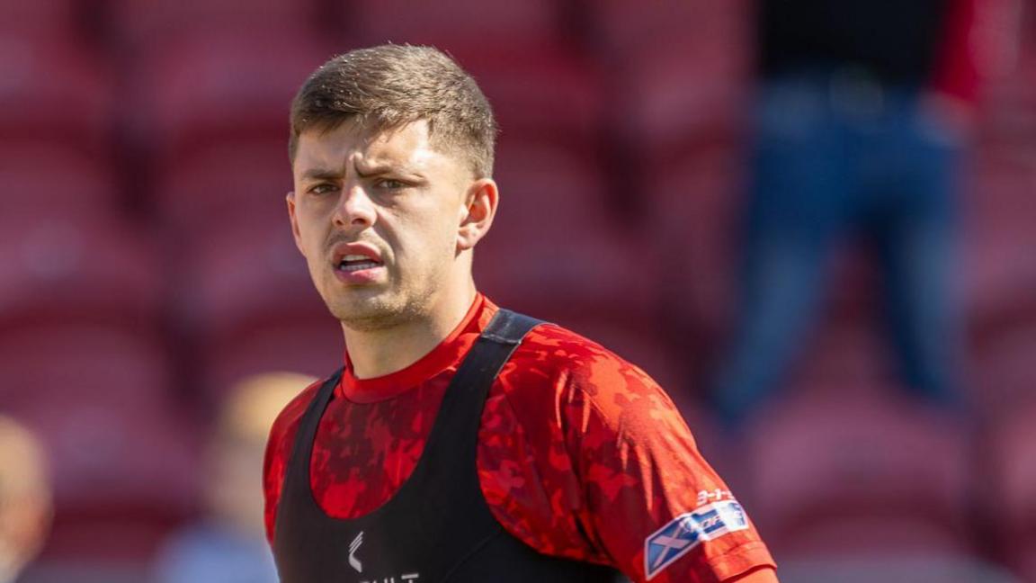 Adam Lewis, who has short, light hair, is pictured wearing a red training shirt and a black training bib over the top of it