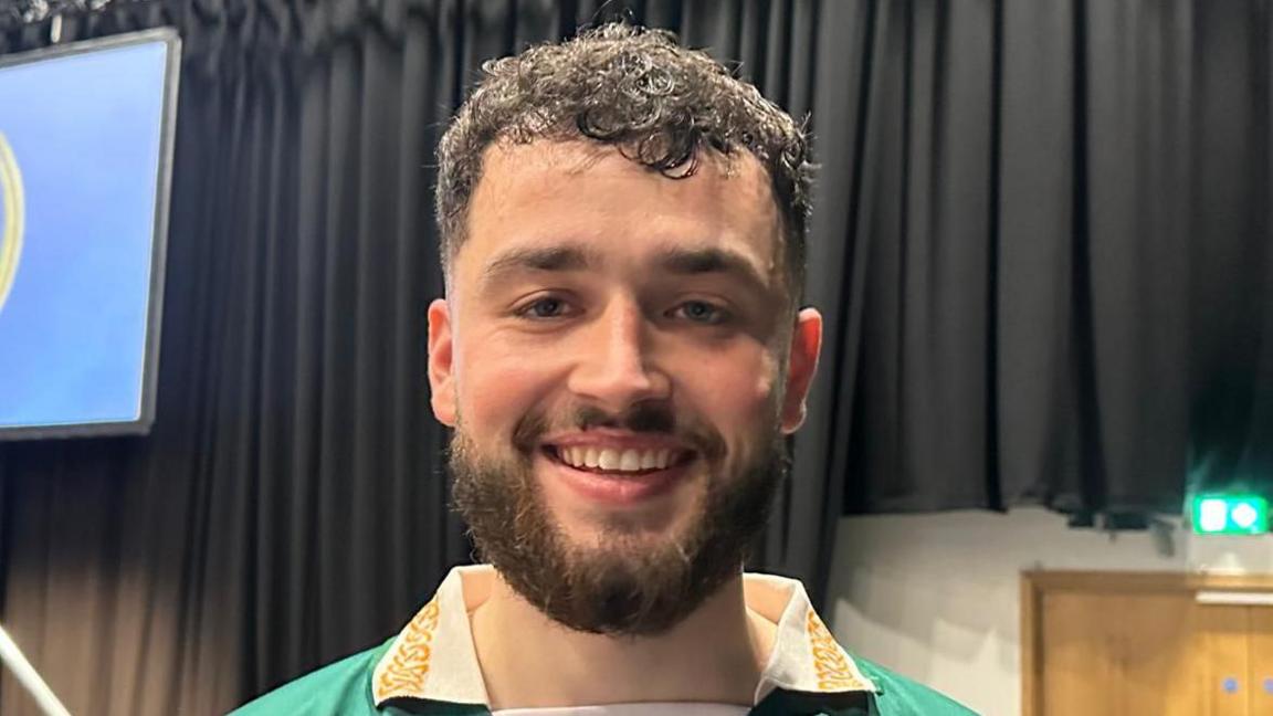Laochra Mac Iomaire has short dark curly hair and a brown beard, and moustache. He is wearing a dark green top with a white and gold collar.