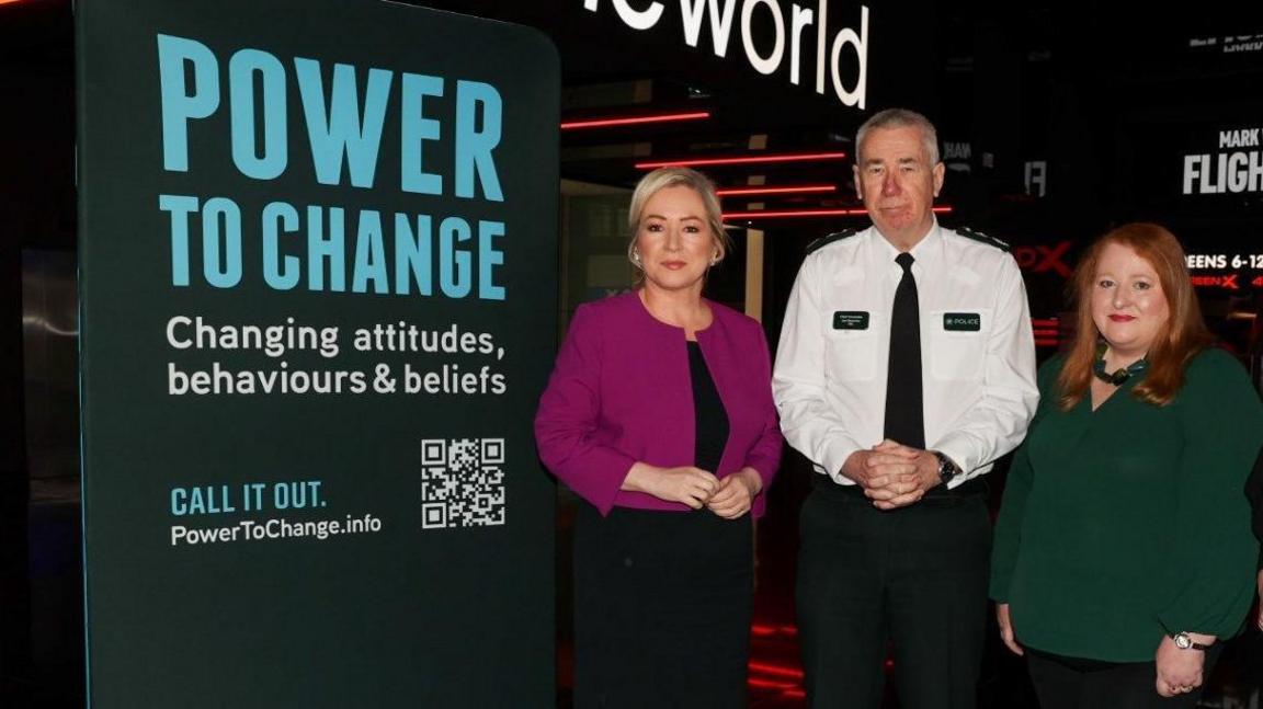 Michelle O'Neill is wearing a black dress and purple/magnolia jacket - her hair is blonde and tied up. 
Jon Boutcher is wearing a PSNI uniform, a white shirt, dark green tie, badges and trousers - his hair is short and white/grey. 
Naomi Long is wearing a green top with black trousers - her hair is red and long. 
All three are standing beside a sign that says 'Power to Change - Changing attitudes, behaviours & beliefs - Call it out' 