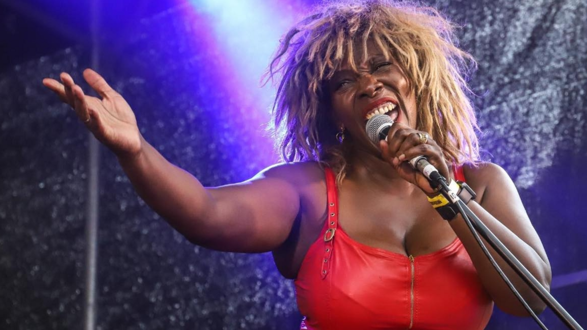 Sass is in a red leather dress and wig to look like Tina Turner. She is singing and has a microphone in her hand close to her face.