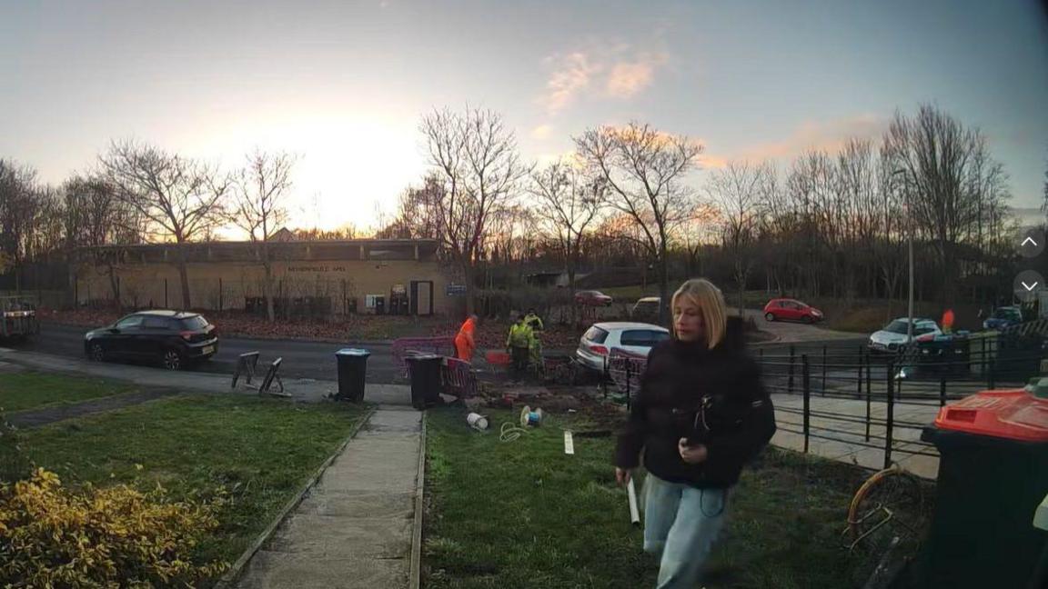 An image captured by a doorbell camera. It shows Melanie Abbott, who has blonde hair and is wearing a padded dark-coloured winter coat, approaching her front door. Behind her, her green front lawn stretches down to the road. At the bottom of the lawn, there are three workers in hi-vis clothing.