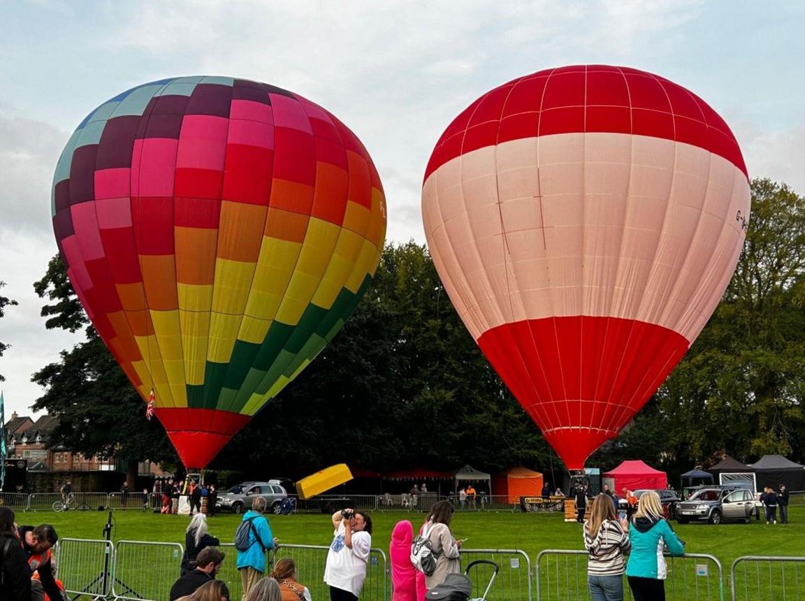 Two hot air balloons on grass, with trees in the background. A dozen people are standing by barriers in the foreground at the bottom of the photo.