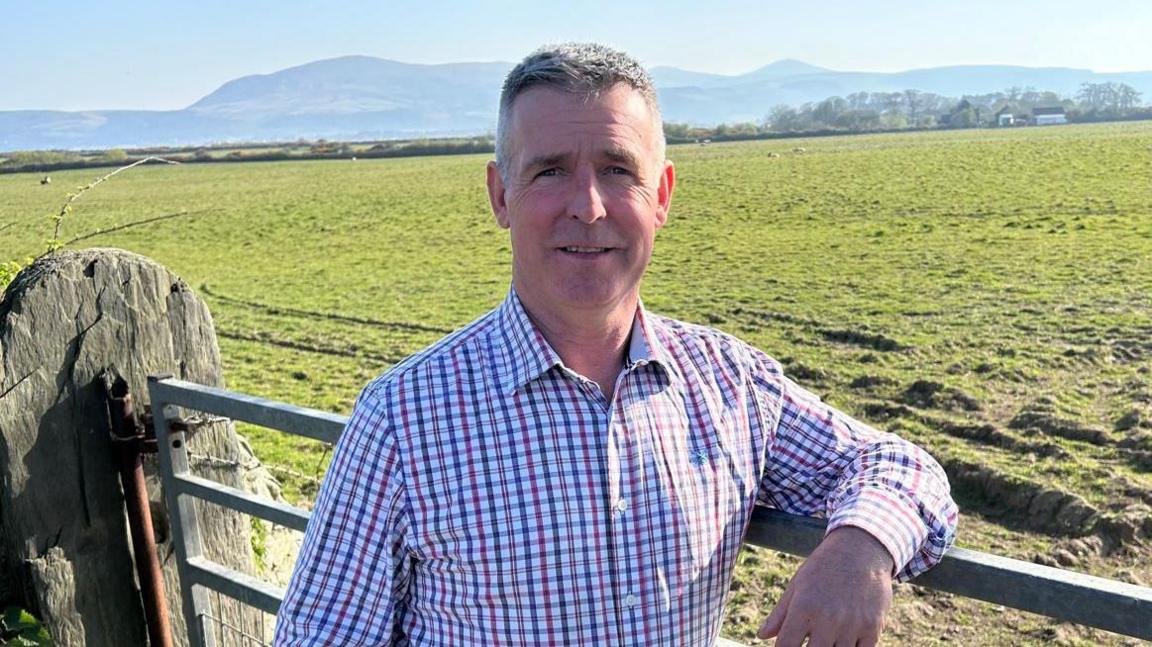 John Teare has short grey hair, and wears a blue and red checked shirt. He leans on a farm gate near a green field.