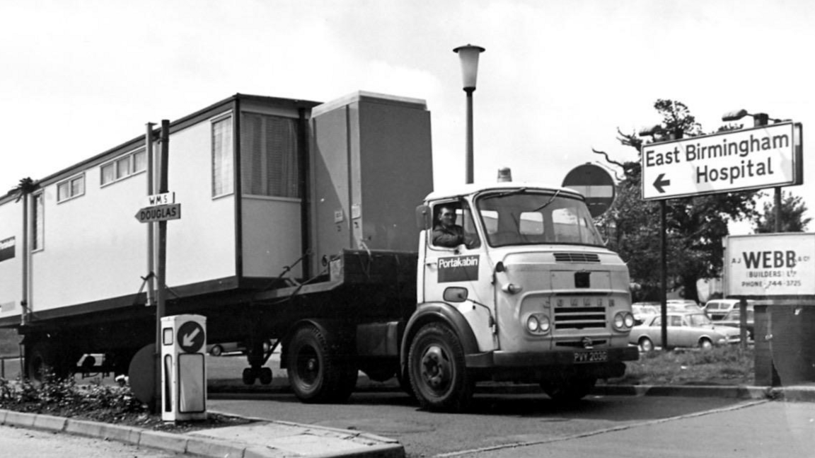 A black and white photo of a lorry with a big building on its back, sitting near a sign which says "East Birmingham Hospital"