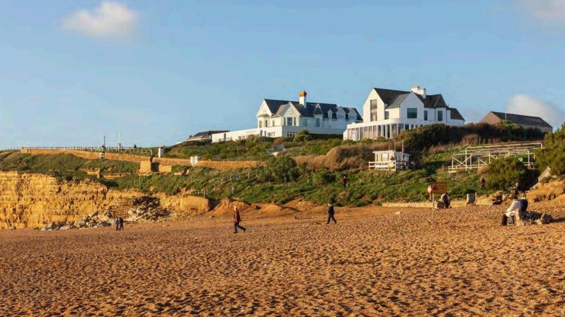 A CGI image of how the finished house would look from the beach. A glass covered extension wraps around the ground floor of the house which stands at the top of a grassy slope. About half a dozen people are walking on the beach in the foreground.