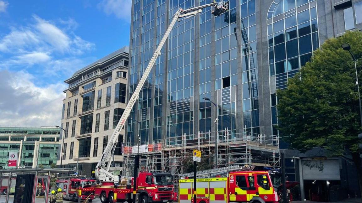A hydraulic aerial platform is by a tall building. Four fire engines are in front of the building on the image.