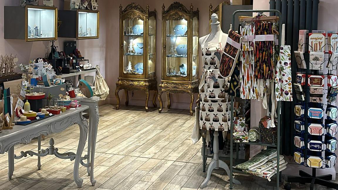 The interior of a shop showing wooden flooring and a table of jewellery and gifts, and some gold display cabinets filled with items in the background