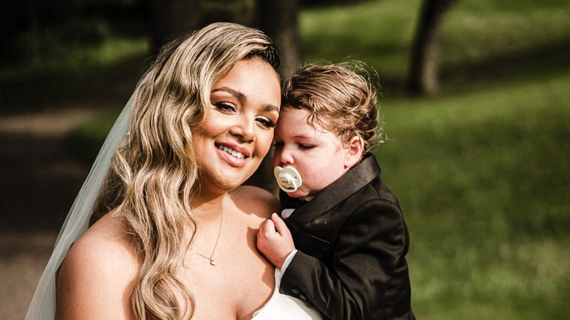 A woman with long blonde hair in a wedding dress with a long white veil is holding her infant/toddler son in her arms. The  boy is wearing a black suit jacket and has a white dummy in his mouth.