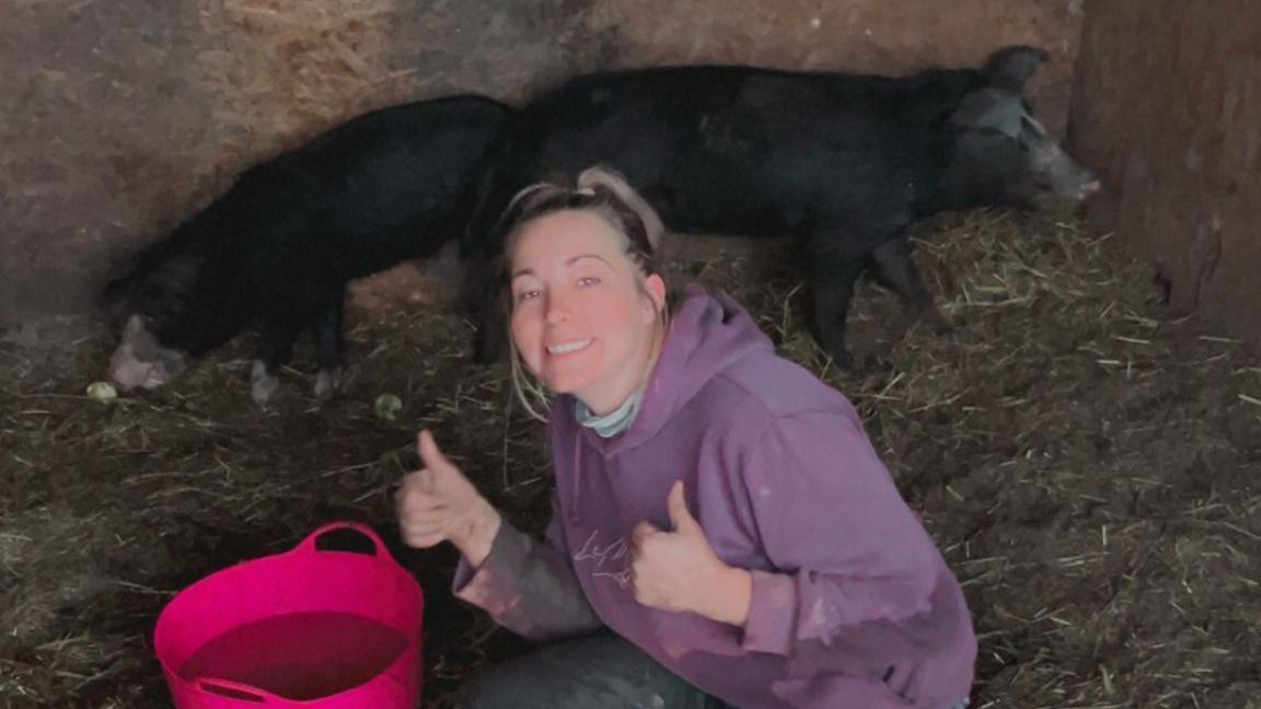 Ms Allen a White woman in her thirties, kneeling down in an animal stables in front of a pink plastic bucket. Behind her are two black pigs. 