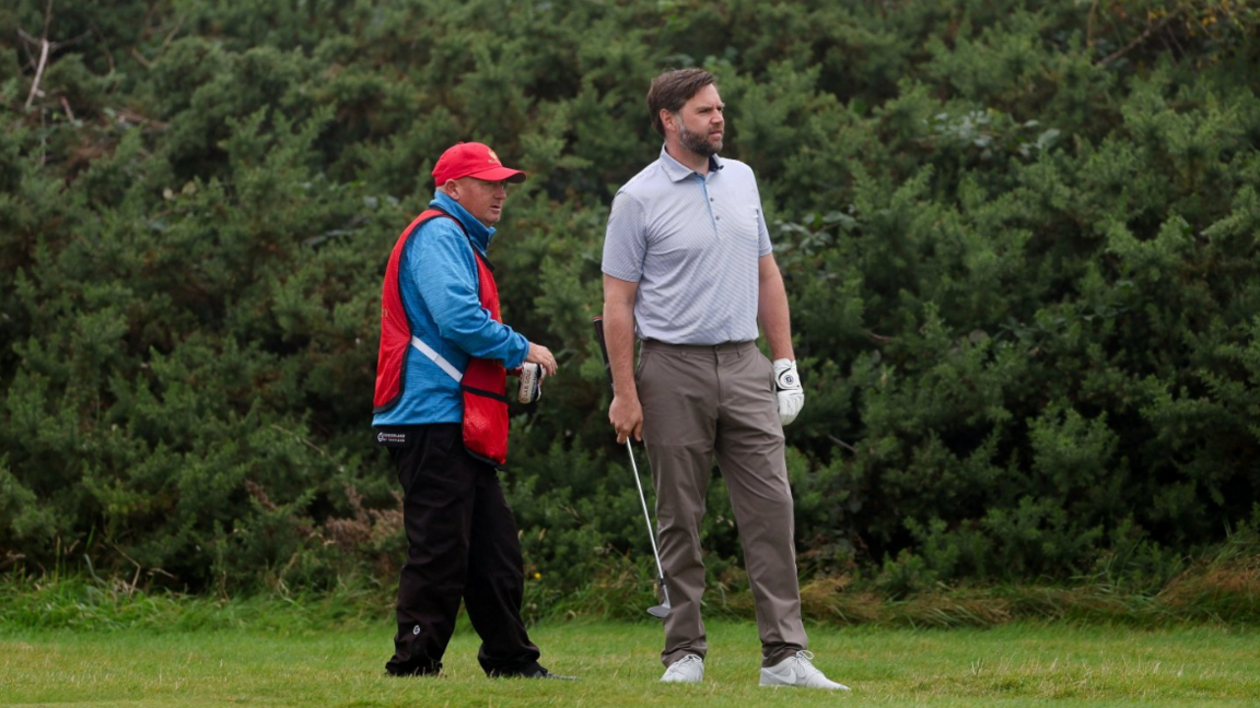 A man with brown hair and bear, wearing a light blue top and brown trousers stands next to a golf caddie wearing a red bib and hat