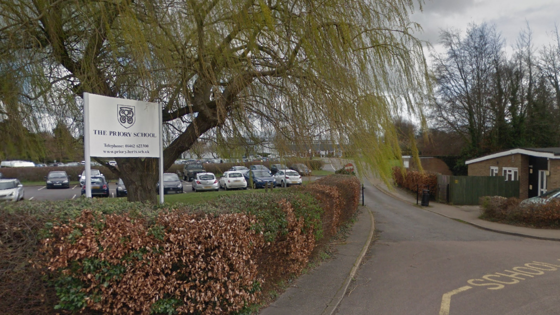 The front gates of Priory School. A large tree stands next to the sign for the school. The car park is beyond that and full of cars. The school itself cannot be seen clearly as it is obstructed by the tree's leaves.