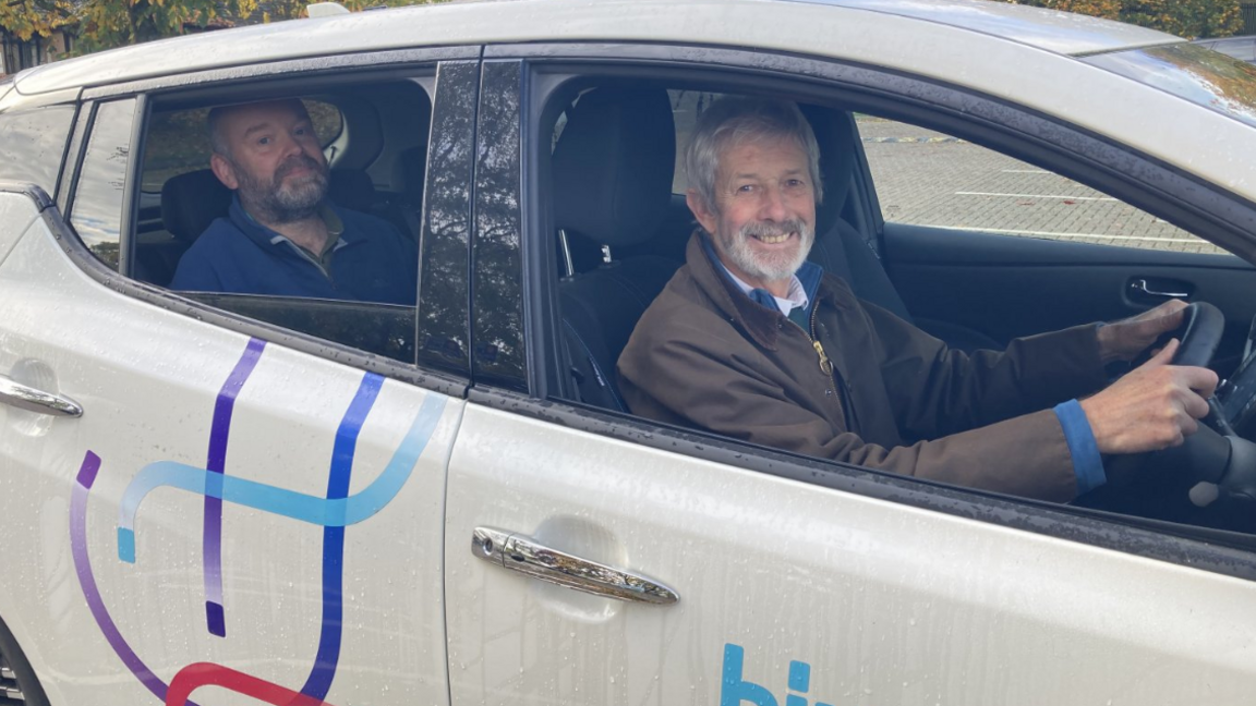Two men seated inside a white car marked with the “hiyacar” logo. The driver is holding the steering wheel, and the passenger is in the back seat. The car is parked outdoors with autumn trees and a paved area visible through the windows.
