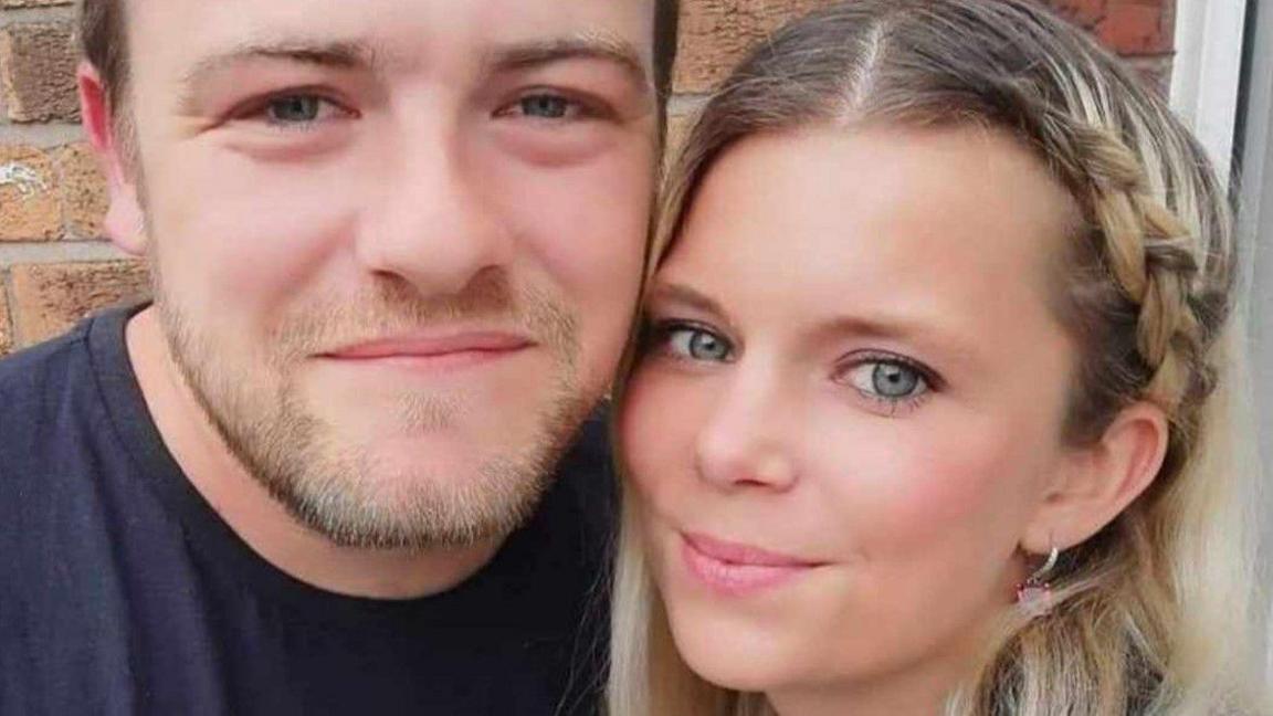 A man and woman look at the camera in front of a brick wall. 