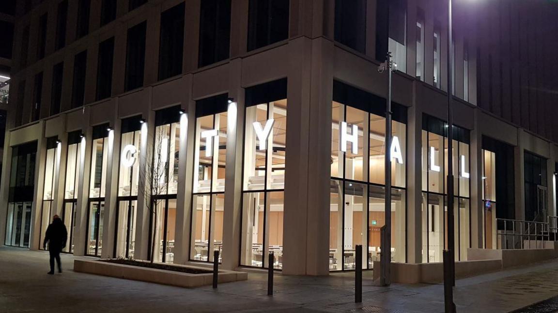 The exterior of Sunderland City Hall at night. Light from the tall building illuminates the street outside. Illuminated letters also spell out: "City Hall."