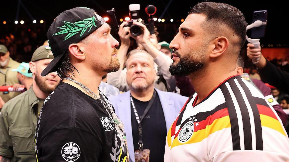 Oleksandr Usyk and Agit Kabayel face off, with Usyk in an IBF-branded black top and Kabayel in a largely white Germany national football team shirt with black, red and gold colours of the country's flag