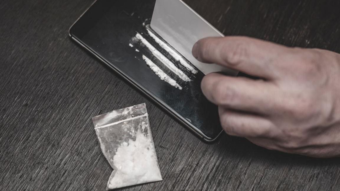 Three lines of white powder on the glass screen of a mobile phone, with a man's hand holding a white plastic bank card. A small snap bag of white powder is lying next to the phone on a table surface.