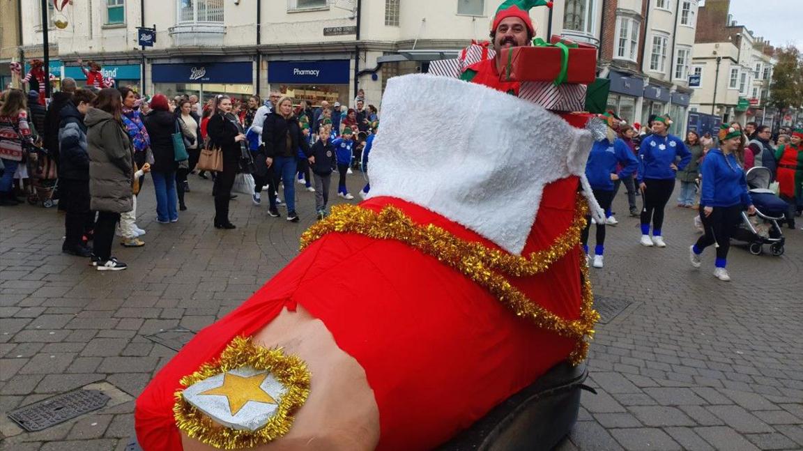 A man dressed as an elf sitting in a giant motorised foot with a red sock on it. His head is poking out of the top of the ankle and he is surrounded by Christmas presents. There is a hole in the toe of the red sock and a large toenail with a gold star on it is poking out.